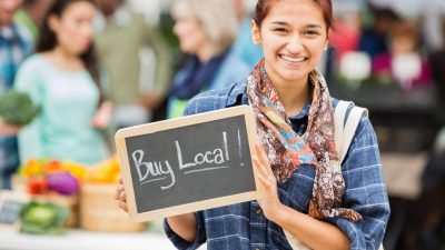 Smiling woman holding a chalkboard sign that says “Buy Local!” at a community market, symbolising support for South Australian small businesses.